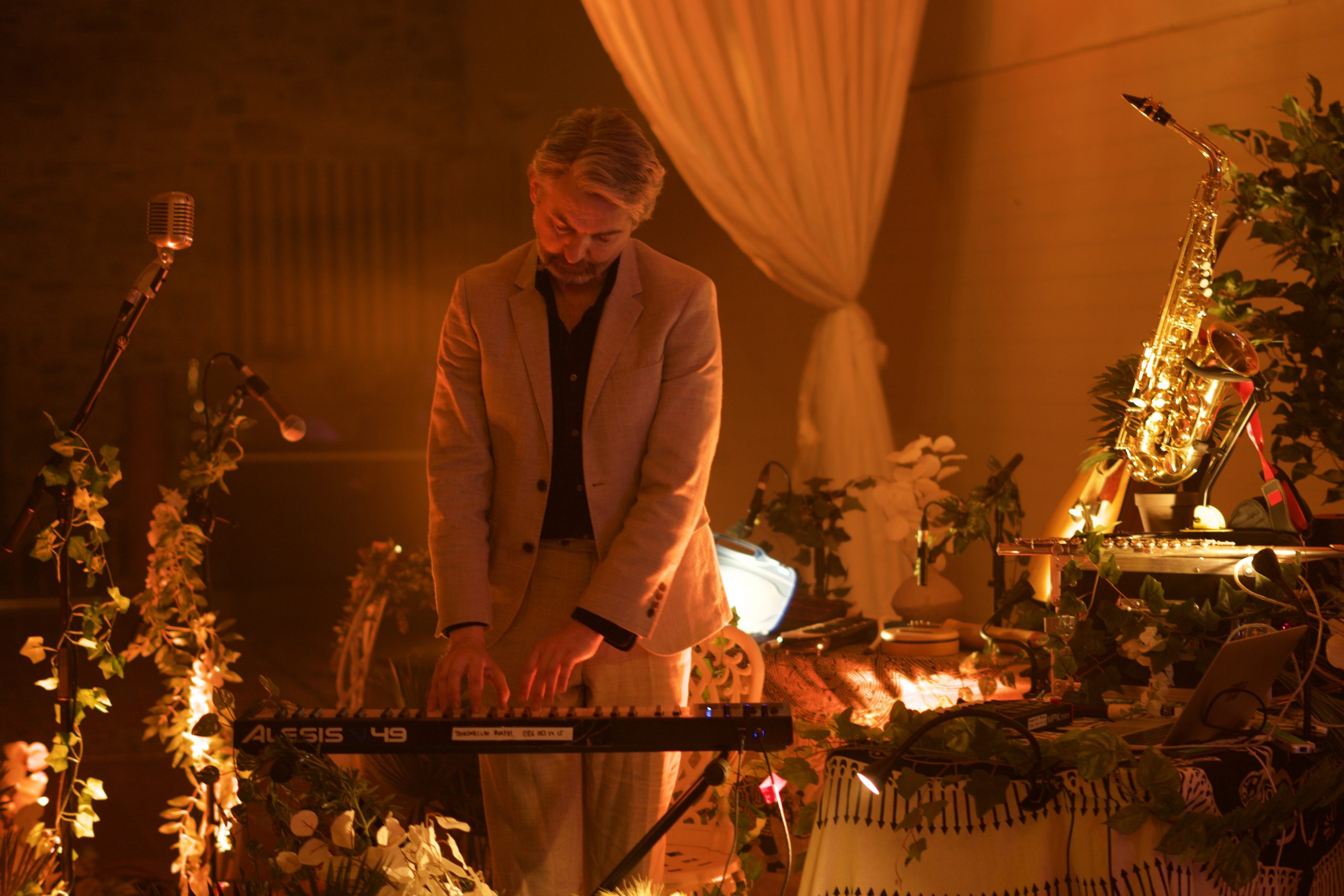 Thunderclap Murphy playing a keyboard in warm amber light, with a saxophone, flowers and draped fabric around him.
