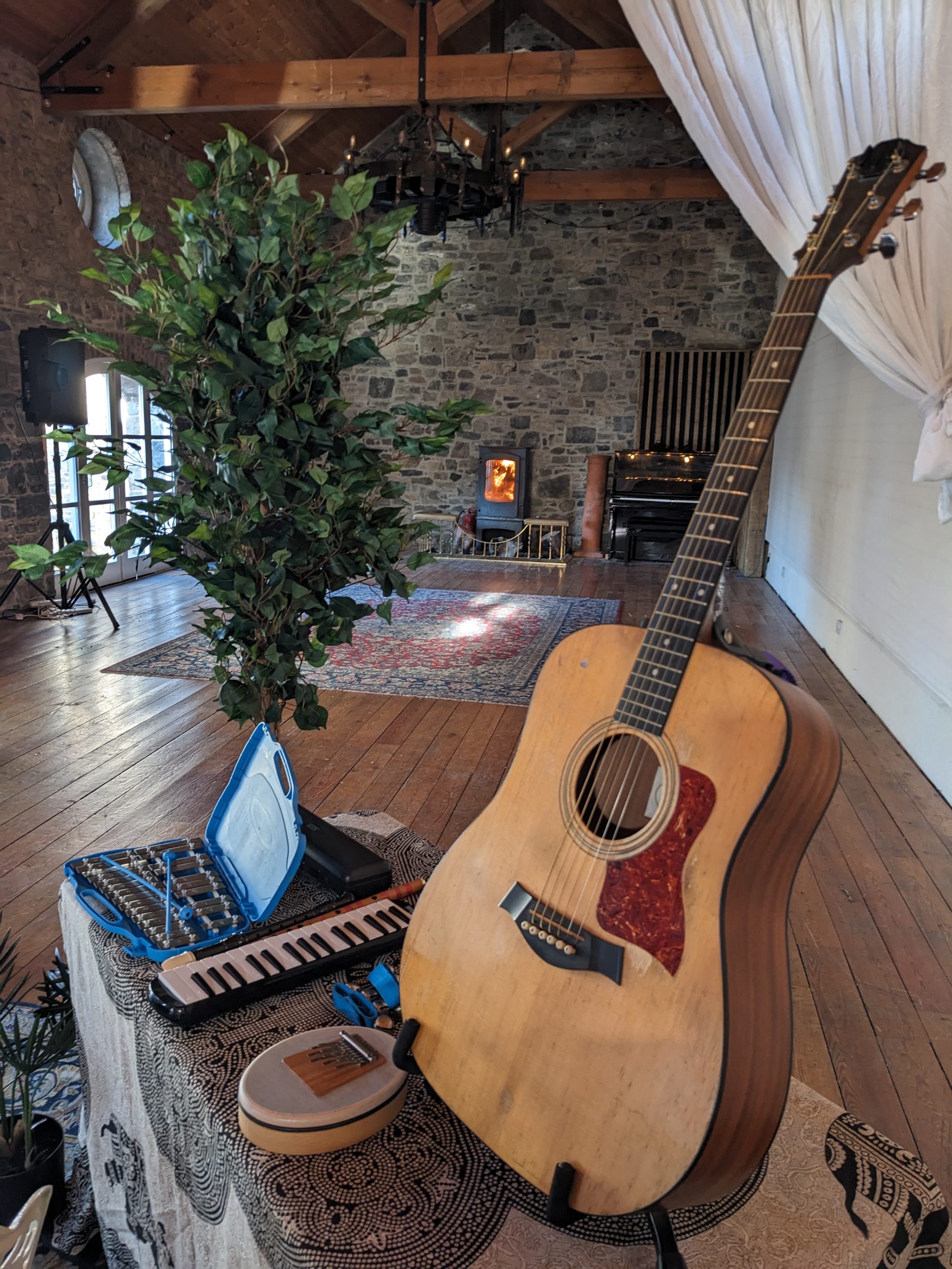 Acoustic guitar, melodica, and hand drum resting in a calm, light-filled room with a wood stove and draped curtains