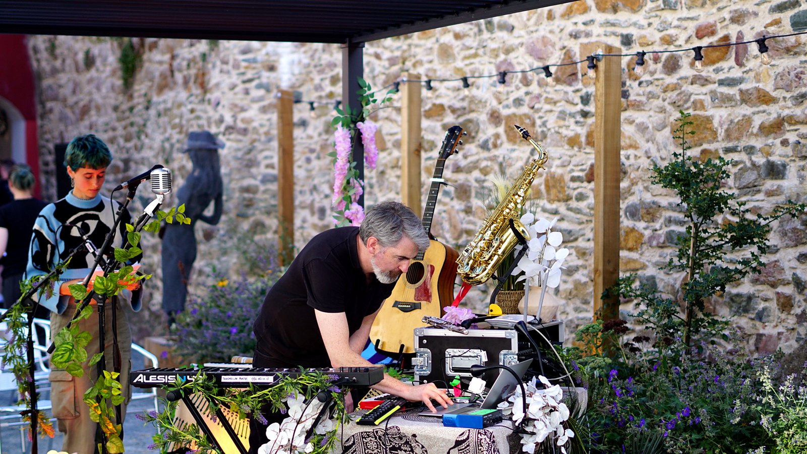 Thunderclap Murphy leaning over the instrument table setting up in the Community Garden before the audience arrived, guitar and saxophone on stands behind, festoon lights above and planting along the stone wall