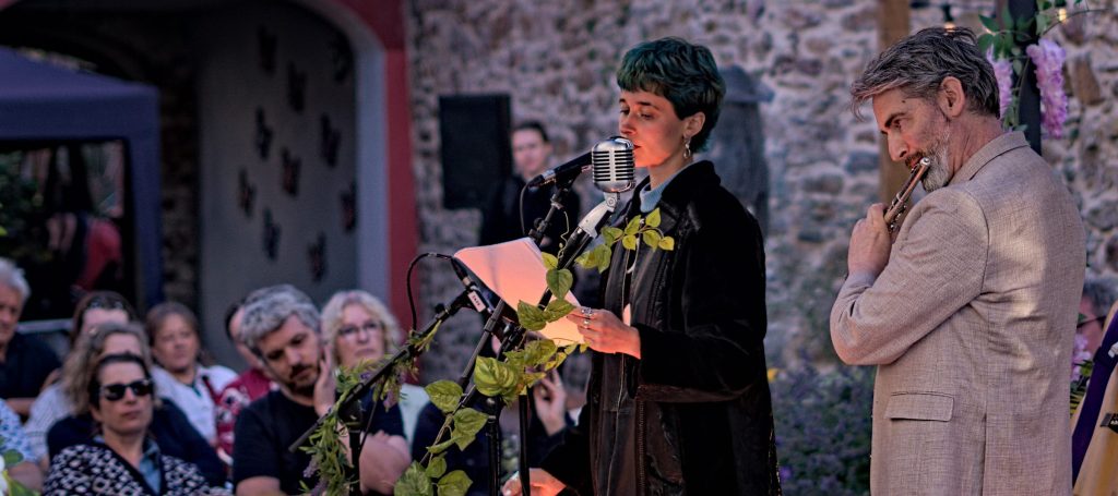 Helen of Troy reading from a page at the microphone while Thunderclap Murphy plays flute beside her, the audience seated in the Community Garden