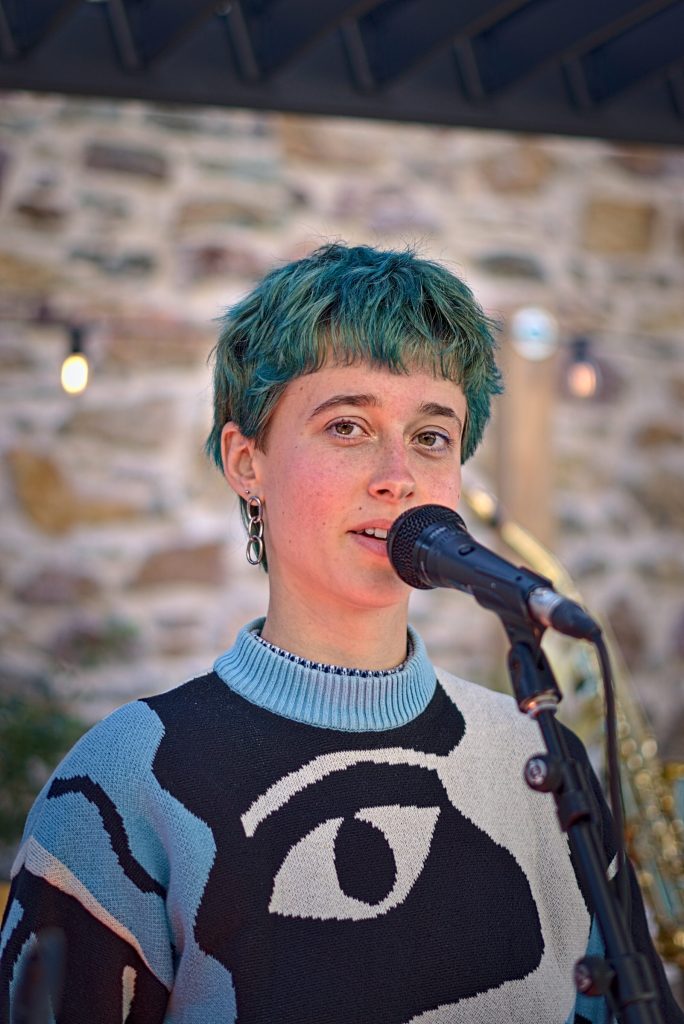 Helen of Troy speaking into a microphone in the Community Garden at Wexford Arts Centre, green hair and a blue and black eye-pattern jumper, stone wall and festoon lights behind