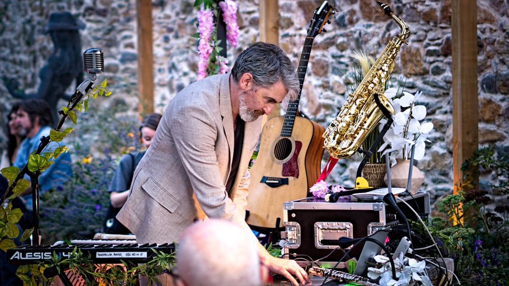 Thunderclap Murphy in a linen jacket leaning over the instrument table in the Community Garden at Wexford Arts Centre, guitar and saxophone on stands against the stone wall, audience at the edges of the frame