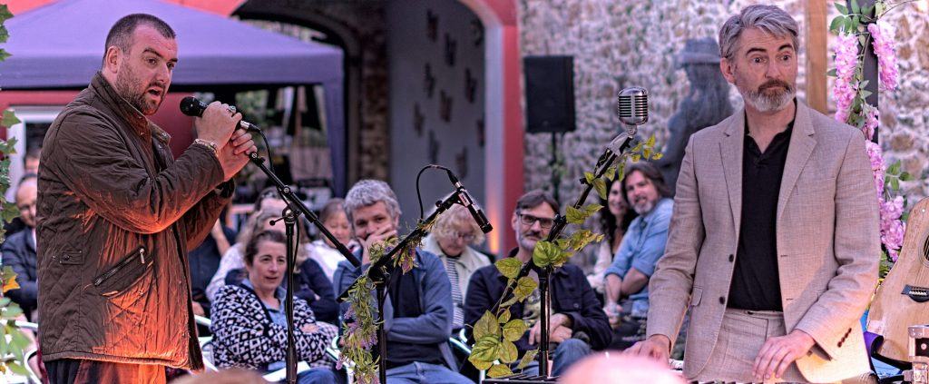 The So Say So host speaking at a microphone on the left while Thunderclap Murphy stands at the keyboard on the right, the audience seated between them