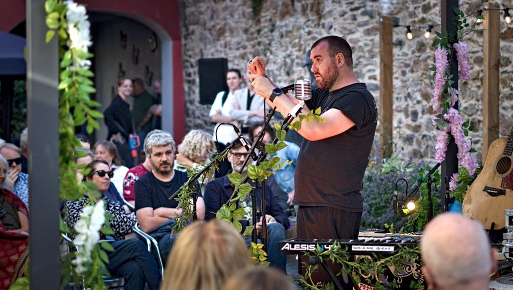 The So Say So host at the microphone in the Community Garden at Wexford Arts Centre, the seated audience beside him and the instruments on stands under the pergola