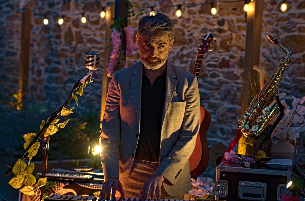 Thunderclap Murphy playing keyboard at dusk under the festoon lights, a vintage ribbon microphone, guitar and saxophone around him against the stone wall