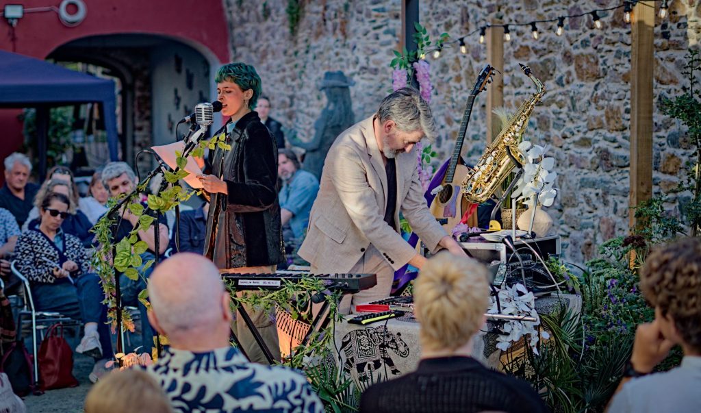 Helen of Troy reading at the microphone while Thunderclap Murphy plays at the instrument table, guitar and saxophone on stands against the stone wall and the audience seated in front