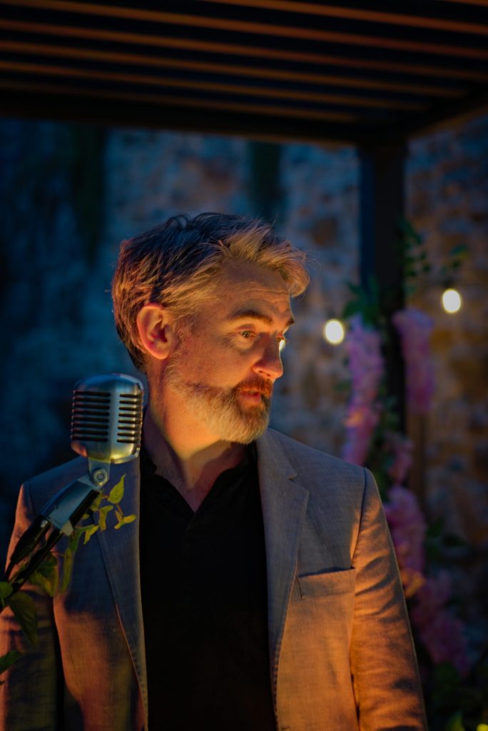 Thunderclap Murphy beside a vintage ribbon microphone at dusk, lit warm against the blue stone wall and pink flowers of the Community Garden