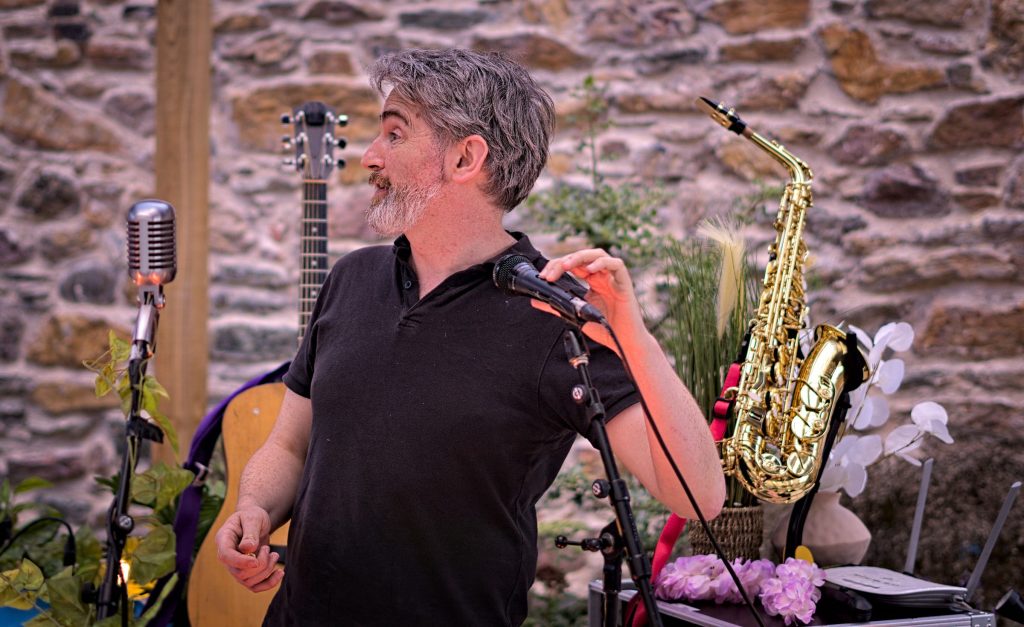 Thunderclap Murphy at the microphone during the soundcheck under the pergola, guitar and saxophone on stands against the stone wall of the Community Garden