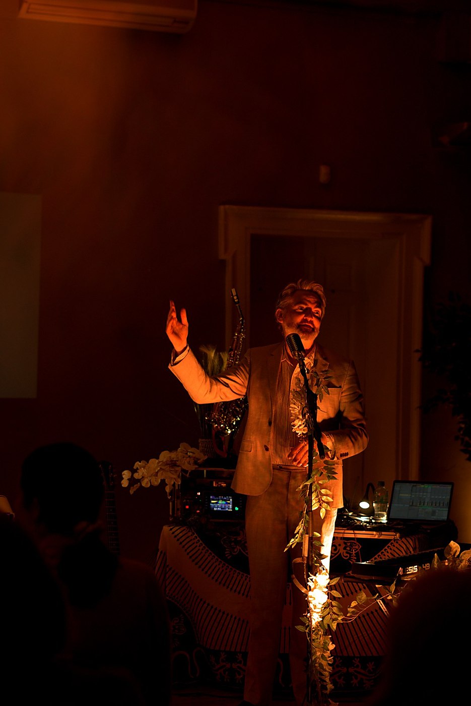 Thunderclap Murphy at the microphone with one hand raised, the keyboard, saxophone and foliage staging behind him