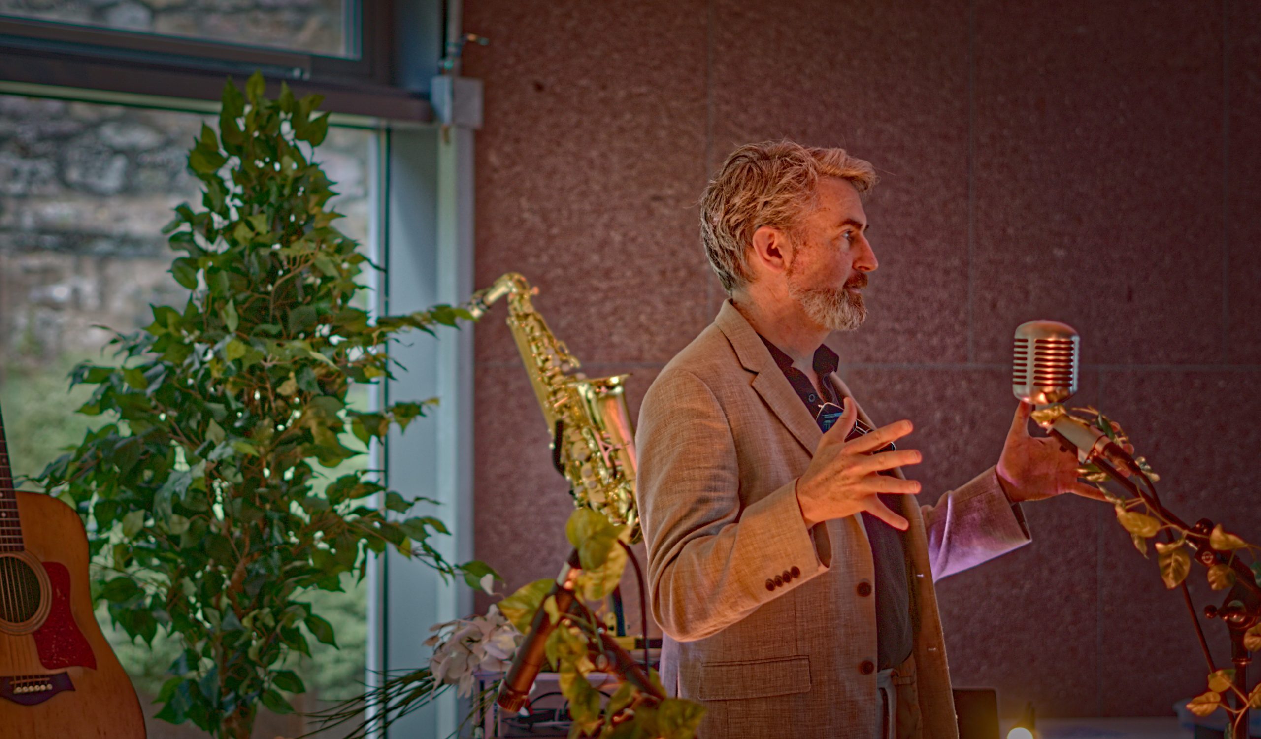 Thunderclap Murphy speaking to the room at a vintage ribbon microphone, a saxophone and guitar behind him.
