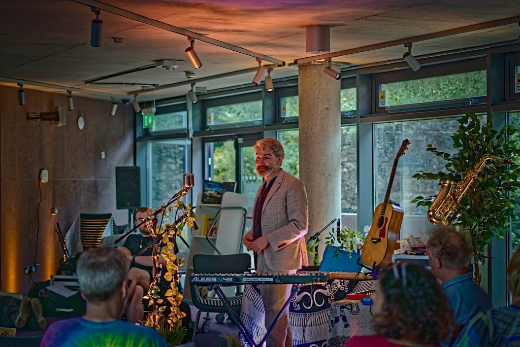 The events room at Wexford Town Library, glazed floor to ceiling on two sides, with Thunderclap Murphy standing at a table of instruments — keyboard, guitar and saxophone — and readers seated in the foreground.